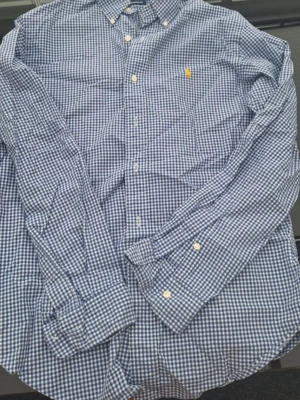 Blårutig skjorta från Ralph Lauren - Klassisk blårutig skjorta från Ralph Lauren i bomull med button-down krage och gul broderad logga på bröstet. Skjortan har lång ärm och vita knappar framtill. Perfekt för en stilren och avslappnad look.