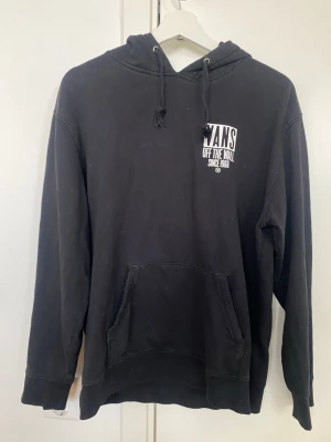 Svart hoodie från Vans med tryck - Svart hoodie från Vans med stor vit logga och texten 'Off The Wall Since 1966' på ryggen och mindre tryck på bröstet. Klassisk känguruficka framtill och justerbar huva med snören. Mjuk bomullskänsla och avslappnad passform. Snören defekta