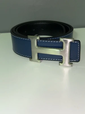 Blått bälte skärp med H-spänne - Snyggt blått skärp med det ikoniska H-spännet i silverfärgad metall. Skärpet har vita sömmar längs kanterna och är gjort i skinn. Perfekt accessoar för att lyfta din outfit.