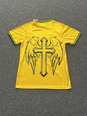 Gul Brasilien t-shirt med Jesusmotiv - Unik gul Brasilien t-shirt från Nike med gröna detaljer på krage och ärmslut. Framsidan har ett stort konstnärligt motiv av Jesus och baksidan pryds av ett kors med vingar. Perfekt för dig som vill sticka ut med en personlig stil.
