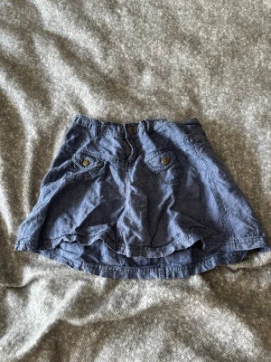 Blå miniskirt med fickor - Snygg blå kortkjol i bomull med två framfickor med knappdetaljer och knappstängning i midjan. Kjolen har en avslappnad passform och är perfekt för varma dagar. Enkel att matcha med olika toppar för en cool vardagslook.