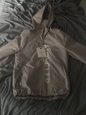 Grå vindjacka från Moncler - Pass formen är lite minde så tror en storlek S skulle kunna ha på sig denna.Snygg grå vindjacka från Moncler med huva och dragkedja framtill. Jackan har stora vita Moncler-bokstäver längs dragkedjan och praktiska fickor med dragkedja. Tillverkad i ett lätt och vindtätt material, perfekt för blåsiga dagar. 