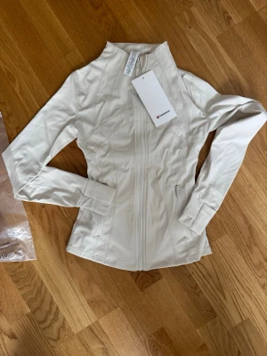 Vit jacka från Lululemon med dragkedja - Snygg vit jacka från Lululemon med figurnära passform och hel dragkedja framtill. Tillverkad i ett stretchigt syntetmaterial och har långa ärmar samt fickor med dragkedja. Perfekt för en sportig och clean look.