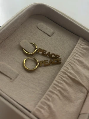 Guldörhängen med PEACE-text - Säljer ett par trendiga örhängen i guldfärgad metall med texten PEACE som hänger från små ringar. Perfekta för dig som vill ha ett statement-smycke med cool vibe. Kommer i en smidig förvaringsask.