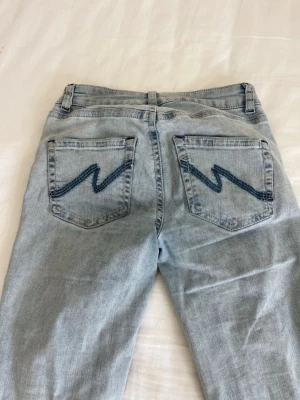 Ljusblå bootcut jeans med slitningar - Säljer ett par ljusblå bootcut jeans med slitna detaljer på knäna och broderade bakfickor med zigzag-mönster. Jeansen har klassisk femficksmodell och är tillverkade i denim. Perfekta för en avslappnad och trendig look.