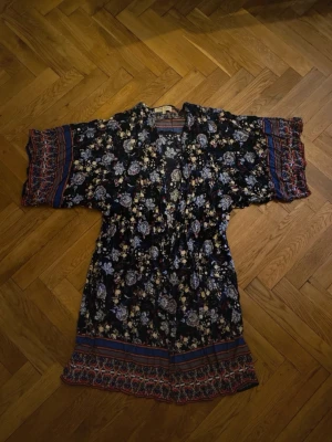 Blommig kimono morgonrock - Snygg morgonrock i kimonomodell med svart botten och blommigt mönster i vitt, lila och gult. Vida ärmar med dekorativa bårder i rött och blått. Lätt och luftig, perfekt för att chilla hemma. Materialet känns mjukt och följsamt.