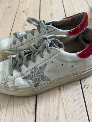 Golden Goose vita sneakers med glitterstjärna - Golden Goose sneakers i vitt läder med grå skosnören, silverglittrig stjärna på sidan och rödglittrig häl. Textdetaljer på sulan och märkeslogga på sidan. Klassisk låg modell med vintagekänsla och robust sula.