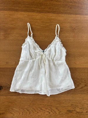 Vit spetslinnetopp från Gina Tricot - Söt vit linnetopp från Gina Tricot med tunna axelband och dekorativ spets. Toppen har broderade blommor och en liten knytning framtill. Perfekt för sommardagar och kan stylas med både jeans och kjol.