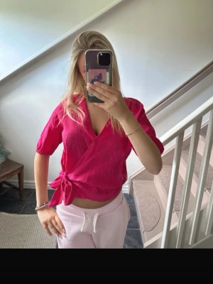 Rosa omlottopp med knyt - Snygg rosa omlottopp med knyt i sidan och korta puffärmar. Toppen har v-ringning och är gjord i ett lätt, strukturerat material som känns skönt mot huden. Perfekt för dig som vill ha en trendig och bekväm look.
