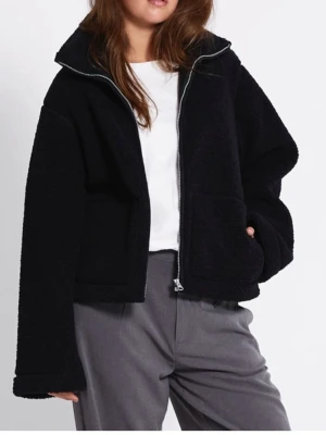 Svart teddyjacka med dragkedja - Mysig svart teddyjacka med hög krage och dragkedja framtill. Jackan har två stora fickor och är tillverkad i fluffigt teddy-material som ger en cool och avslappnad look. Perfekt för att hålla sig varm under kyliga dagar.