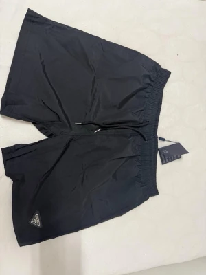 Svarta shorts från Prada med snörning - Säljer ett par svarta shorts från Prada med elastisk midja och snörning. Shortsen har en klassisk passform och är tillverkade i 100% nylon. Diskret Prada-logga på benet och stilren design som passar perfekt till sommaren.