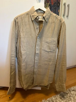 Beige linneskjorta från Gant - Stilren beige skjorta från Gant i linne med klassisk krage och knappstängning framtill. Skjortan har en bröstficka och långa ärmar med knappar vid manschetterna. Perfekt för en avslappnad men snygg look.