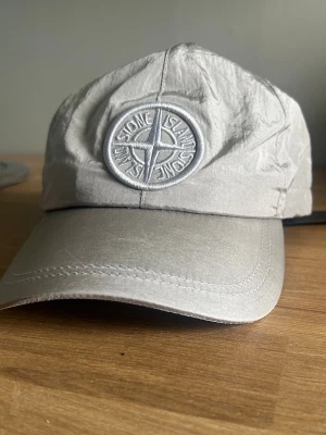 Keps från Stone Island - Snygg ljusgrå keps från Stone Island med det klassiska kompassloggan broderad framtill. Kepsen har böjd skärm och justerbar rem bak. Tillverkad i ett lätt och slitstarkt material som ger en clean och modern look.