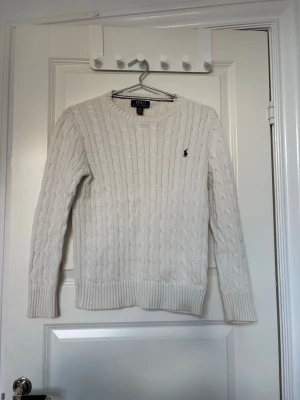 Vit kabelstickad tröja Polo Ralph Lauren - Kabelstickad benvit tröja från Polo Ralph Lauren med rund halsringning och den klassiska broderade loggan i marinblått på bröstet. Tröjan har långa ärmar och ribbade muddar vid ärmslut och nederkant. Perfekt för en clean och stilren look. Tröjan inköpt år 2025, äkta! Ralph Lauren 10-12 är för 10-12år. Köparen betalar frakt. 
