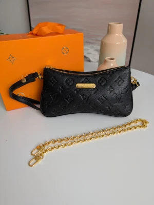 Iouis Vuitton Pochette Liv 🖤 - Sofistikerad axelremsväska i svart präglat läder med guldkedja Förhöj din stil med en tidlös och mångsidig afton- och vardagsväska i premiumutförande. Denna eleganta modell har en modern, svagt kurvad silhuett och erbjuder en perfekt balans mellan klassisk lyx och minimalistisk finess. Detaljer och egenskaper: Material & Struktur: Tillverkad i högkvalitativt, fylligt svart läder med en distinkt kornig textur där det ikoniska monogrammönstret är sofistikerat präglat i materialet. 