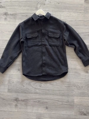 Mörkgrå overshirt med fickor - ENDAST TESTAT! Stilren mörkgrå overshirt med klassisk krage och två stora bröstfickor med lock och knapp. Rak passform och knäppning framtill. Perfekt att bära som lager över t-shirt eller hoodie. Materialet ser ut att vara mjukt och lite tjockare, troligen ullblandning. Prislapp medföljer! Tyvärr inget kvitto.
