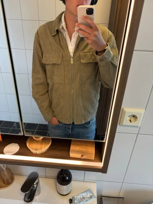 Beige manchester overshirt med dragkedja - Säljer en beige overshirt i manchestertyg med dragkedja framtill och två stora bröstfickor. Overshirten har en avslappnad passform och klassisk krage, perfekt att slänga över en t-shirt eller skjorta. Materialet är mjukt och ger en retrovibe.