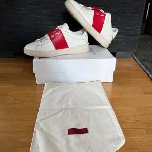 Valentino Open  - Säljer ett par vita Valentino Open sneakers med bred röd rem över mitten. Skorna har rund tå, platt sula och vita snören. Klassisk och stilren design med diskret Valentino-logga på plösen. Kommer med originalkartong och dustbag. Skorna är inte tvättade på bilden och går att göra finare✅✅ vid frågor och funderingar är det bara att skriva till mig 🤩🤩 skorna är 44 men passar till mig som har 42/43 opens sitter väldigt smått så skulle säga att de är runt 42/43 i storleken ✅ kom med prisförslag🤩🤩