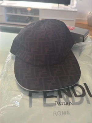 Svart keps med Fendi-logga - Säljer en svart keps från Fendi med det klassiska FF-monogrammet över hela kepsen. Kepsen har böjd skärm och är tillverkad i ett tygmaterial som känns lyxigt och stilrent. Perfekt för dig som vill ha en exklusiv accessoar med tydlig logga.