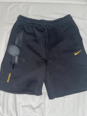 Svarta NOCTA x Nike shorts - Svarta shorts från NOCTA x Nike med gul logga och detaljer. Mjuk bomullsmix, dragsko i midjan och dragkedjeförsedd ficka på sidan. Snygga broderade detaljer bak och framtill. Perfekta för en avslappnad och sportig stil.