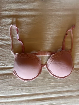 Ljusrosa push-up bh med spets - Söt ljusrosa push-up bh med vadderade kupor och fina spetsdetaljer längs kanten. Justerbara axelband och hakknäppning bak. Materialet är mjukt och bekvämt, perfekt för dig som vill ha både stöd och en feminin look.