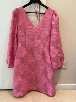 Rosa kort klänning med puffärmar - Supersöt rosa kort klänning med strukturmönster och v-ringning både fram och bak. Klänningen har långa puffärmar och knytband i nacken för extra detalj. Perfekt för dig som vill sticka ut med en färgglad och trendig look.
