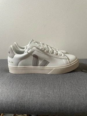 Veja skor - Stilrena vita sneakers från Veja med grå V-logga på sidan och grå hälflik. Skorna har klassisk låg profil, vita snören och sula i offwhite. Tillverkade i läder och mocka för en clean och modern look.