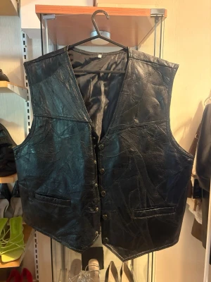 Svart skinnväst med knappar, size XXL/44 - Snygg svart väst i patchwork-liknande skinnimitation med knäppning framtill och två fickor. Västen har en klassisk V-ringning och är helfodrad för extra komfort. Perfekt för dig som vill addera en edgy touch till din outfit.