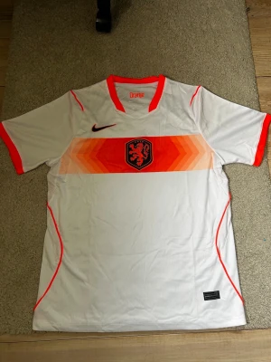 Vm Nederländerna Nike fotbollströja - Säljer en vit fotbollströja från Nike med Nederländernas landslagsemblem (KNVB) på bröstet. Tröjan har orange detaljer, V-ringning och korta ärmar. Tillverkad i lätt och ventilerande material, perfekt för träning eller match.