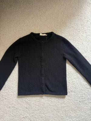 Svart klassisk kofta med rund hals - En stilren svart kofta med rund halsringning och knäppning framtill. Koftan har långa ärmar och en rak passform, perfekt att slänga över en t-shirt eller blus. Materialet är mjukt och lite stickat, vilket ger en mysig känsla.