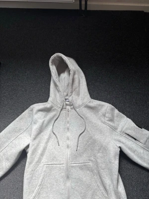 Ljusgrå zip hoodie med fickor - En nästan helt ny stilren ljusgrå cp company hoodie med dragkedja framtill och två stora magfickor. Hoodien har justerbar huva med snören och ribbade muddar vid ärmar och nederkant. Perfekt för en avslappnad look och enkel att matcha med andra plagg.