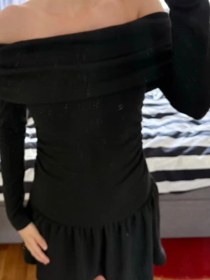 Svart offshoulder klänning - Trendig svart klänning med offshoulder-design och långa ärmar. Klänningen har en åtsittande passform upptill och en lätt volang vid kjolkanten. Perfekt för dig som gillar en stilren och elegant look.