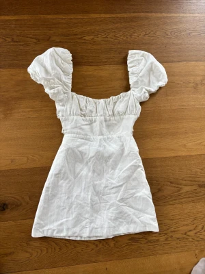 Vit kort puffärmad klänning Zara - Supersöt vit kort klänning från Zara med puffiga korta ärmar och rynkade detaljer upptill. Klänningen har en feminin siluett och är tillverkad i ett lätt tyg som passar perfekt till varma dagar. Enkel att styla med sneakers eller sandaler.