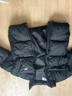 Svart pufferjacka från Nike - Snygg svart pufferjacka från Nike med diskret logga på bröstet. Jackan har hög krage, dragkedja framtill och breda, vadderade paneler som ger en cool och sportig look. väldigt varm och tjock puffer jacka perfekt för kalla vinterdagar och enkel att matcha med olika outfits. 