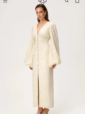 Lång ljus gul Adoore  klänning  - Elegant långklänning i ljus färg med v-ringning och dekorativa knappar framtill. Klänningen har voluminösa långärmar och ett subtilt blommigt mönster i tyget. Tillverkad i ett glansigt material som ger en exklusiv känsla.