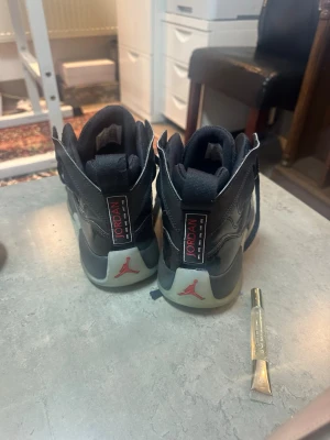 Svarta Air Jordan sneakers - Säljer ett par svarta Air Jordan sneakers med högt skaft och klassisk Jumpman-logga i rött på hälen. Skorna har detaljer i vitt och grått samt tydliga Jordan-märken på baksidan. Perfekta för dig som gillar streetwear och sportig stil.