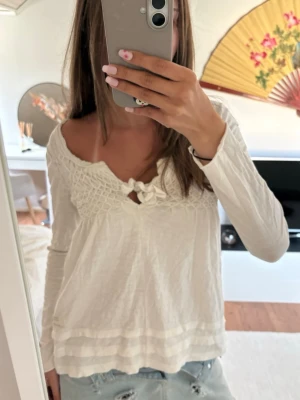 Vit långärmad blus med rosett - Söt vit blus med lång ärm och smockad överdel. Toppen har en v-ringning med en liten rosett framtill och dekorativa sömmar längst ner. Perfekt till jeans eller kjol för en avslappnad look.