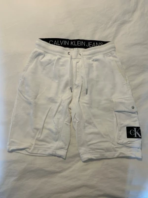 Vita Calvin Klein shorts - Snygga vita shorts från Calvin Klein Jeans med resårmidja och snörning. Shortsen har en ficka på sidan med CK-logga och är tillverkade i mjuk bomull. Perfekta för sommaren och har en avslappnad passform.