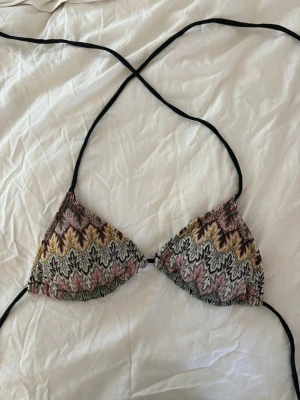 Mönstrad trekantsbikini från Design By Si - Säljer en snygg trekantsbikini från Design By Si med färgglatt, vågigt mönster i svart, rosa, gul och grön. Bikinin har tunna svarta knytband och klassisk trekantsform. Perfekt för stranden eller poolen i sommar! Ifall att ni vill ha bättre bevis av märke, kan jag skicka bild av nedredelen var det finns märket fast.