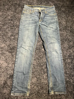 blå raka jeans från Levi's - Säljer ett par klassiska ljusblå jeans från Levi's med raka ben och snygg slitning. Jeansen har fem fickor, gylf med dragkedja och knapp samt gul kontrastsöm. Tillverkade i bomull för en skön och avslappnad look. Storlek 30/32