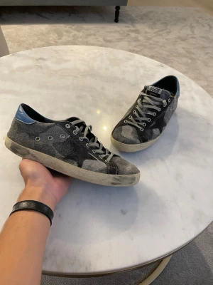 Golden Goose skor - Säljer ett par grå sneakers från Golden Goose med cool patchwork-design och vita snören. Skorna har detaljer i mocka och textil samt en blå hälflik. Klassisk GGDB-logga på plösen och slitna sulor för en trendig look.