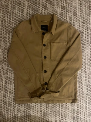 Beige overshirt från Jack & Jones - Stilren beige overshirt från Jack & Jones i bomullstyg. Klassisk krage, svarta knappar framtill och en bröstficka. Perfekt lager-på-lager-plagg med enkel design som funkar till många outfits.