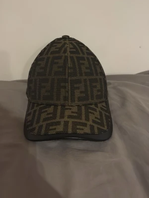  med FF-mönster - Snygg  keps med all-over FF-logga i svart och olivgrönt. Klassisk böjd skärm med svart kant och justerbar baksida. Materialet ser ut att vara en mix av tyg och eventuellt konstläder på skärmens kant. Perfekt för dig som gillar streetwear och loggor.