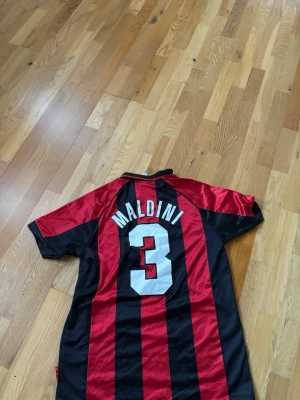 AC Milan fotbollströja Maldini #3 - Säljer en klassisk AC Milan matchtröja från Adidas med Maldini och nummer 3 på ryggen. Tröjan har röda och svarta vertikala ränder, Champions League-märke på ärmen och Opel som sponsor på bröstet. Perfekt för dig som älskar fotboll och retrostil.