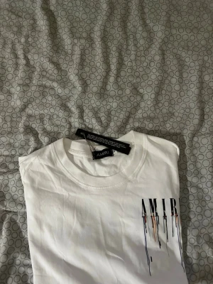 Vit AMIRI t-shirt med färgstänk - Säljer en vit t-shirt från AMIRI med tryck på ryggen där det står AMIRI och färgstänk i svart, orange och blått som rinner ner. T-shirten har rund hals och korta ärmar. Perfekt för dig som gillar streetwear och vill sticka ut.