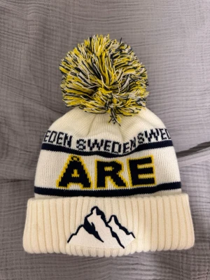 Vit Åre mössa med tofs och brodyr - Säljer en vit stickad mössa med stor tofs i vitt, gult och blått. Mössan har texten 'ÅRE' i gult och blått samt 'SWEDEN' runt om. Broderat bergmotiv framtill och uppvikt kant. Perfekt för kalla dagar i fjällen.