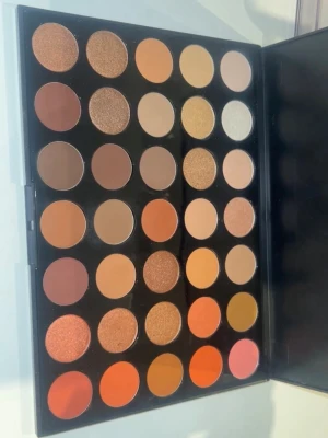 Stor ögonskuggspalett i varma toner -  ögonskuggspalett med 35 nyanser i varma färger som brunt, guld, orange och nude. Paletten innehåller både matta och skimrande skuggor i rund form, perfekt för att skapa olika ögonlooks. Förpackningen är svart och stilren.