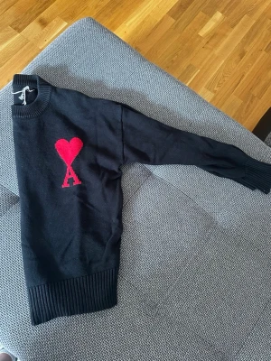 Svart stickad ami Paris - Svart stickad tröja med ett stort rött hjärta och bokstaven A på bröstet. Tröjan har ribbstickade muddar vid ärmslut och nederkant samt rund halsringning. Materialet ser ut att vara mjukt och bekvämt, perfekt för kyligare dagar.