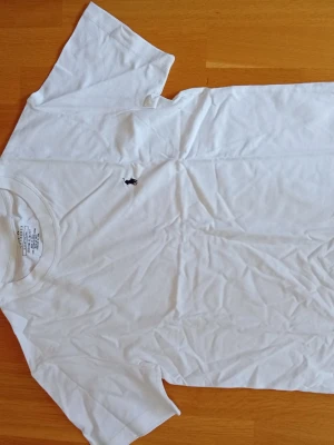 Vit t-shirt med broderad logga - Klassisk vit t-shirt i bomull med korta ärmar och rund hals. På bröstet finns en liten svart broderad logga med en ryttare. Perfekt basplagg som funkar till allt och har en clean look.