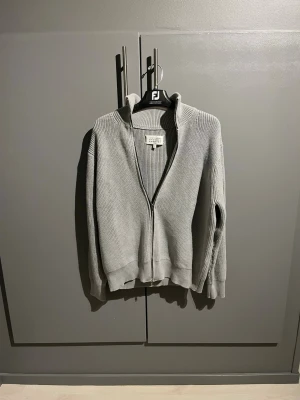 Maison Margiela ziptröja - Ljusgrå stickad tröja med dragkedja framtill och hög krage. Tröjan har ribbade muddar vid ärmslut och nederkant samt en stilren, enkel design. Perfekt för lager på lager och passar till det mesta.